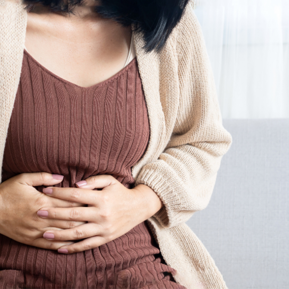 Abnormal Bleeding Woman seated on a sofa in a brown dress and cream cardigan, resting both hands on her lower abdomen